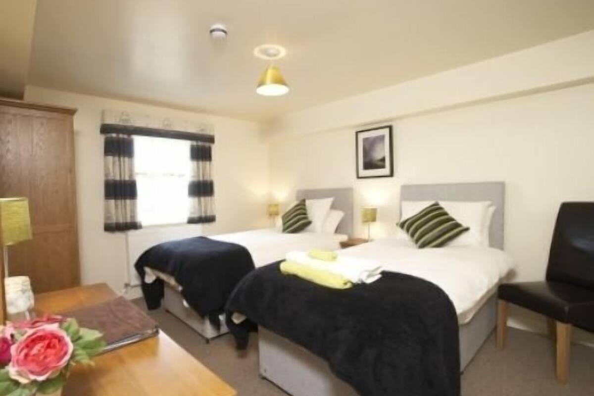 A hotel room with two beds, a desk, a chair, a window with curtains, a picture on the wall, and a vase with flowers.