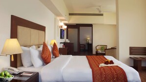 A hotel room with a large bed, a bedside table with a lamp, a phone, and a bowl of fruit. There is a desk with a chair and a small table with a lamp. - Legend Sarovar Portico Baddi (Baddi)