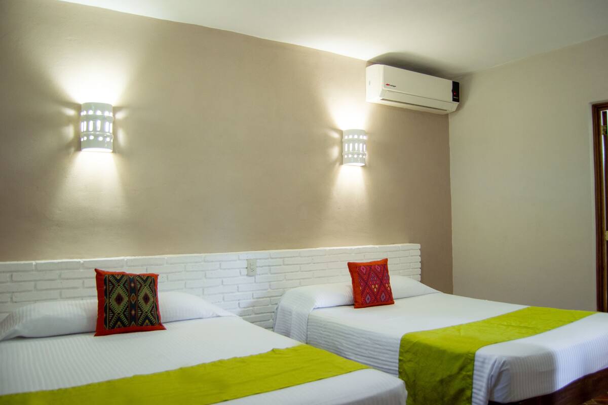 Une chambre d’hôtel avec deux lits, chacun doté d’une tête de lit blanche et d’une couverture verte. Deux lampes sont fixées au mur et un appareil de climatisation est installé au-dessus des lits.