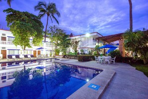 A swimming pool with lounge chairs and umbrellas, surrounded by palm trees and a building with multiple balconies.
