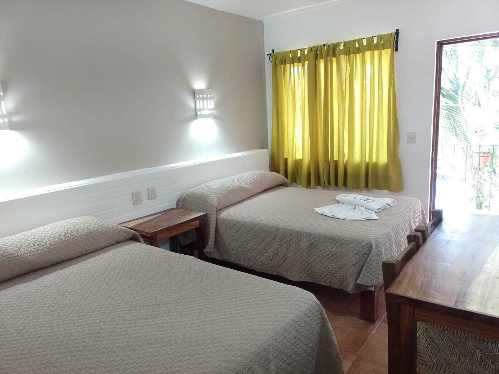 A hotel room with two beds, a wooden nightstand, a small table, and a window with yellow curtains.
