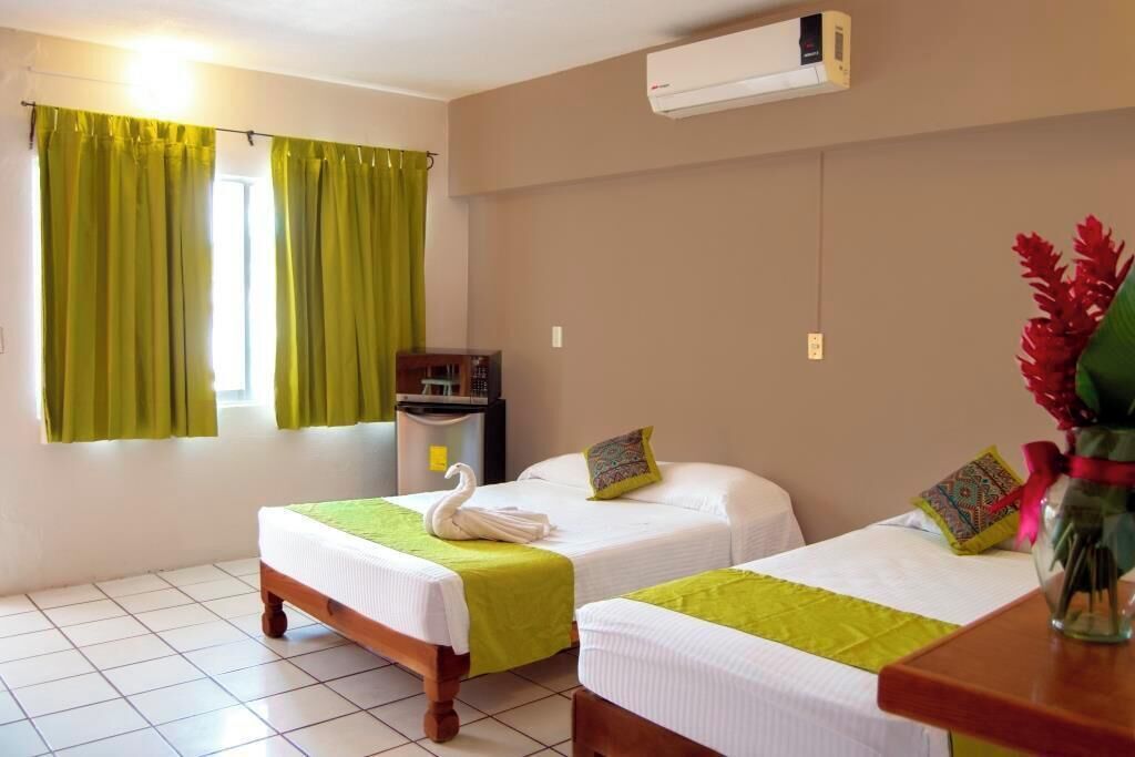 A hotel room with two single beds, a small desk, an air conditioner, and a window with green curtains.