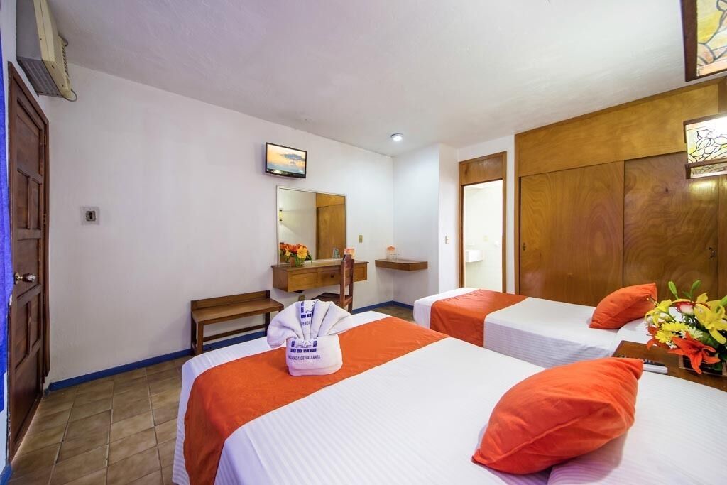 A hotel room with two beds, a wooden wardrobe, a bench, a mirror, and a small table with a vase of flowers.