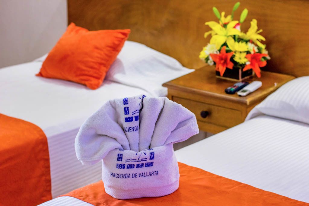 Una habitación de hotel con una cama bien hecha, una mesita de noche de madera con un jarrón de flores y una toalla con texto.