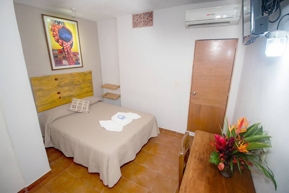 Une petite chambre propre, avec un lit, une tête de lit en bois, une armoire en bois, une petite table sur laquelle est posé un vase de fleurs, et un appareil de climatisation fixé au mur.
