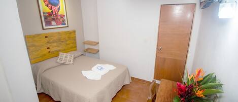 A small, clean room with a bed, a wooden headboard, a wooden wardrobe, a small table with a vase of flowers, and an air conditioning unit on the wall.
