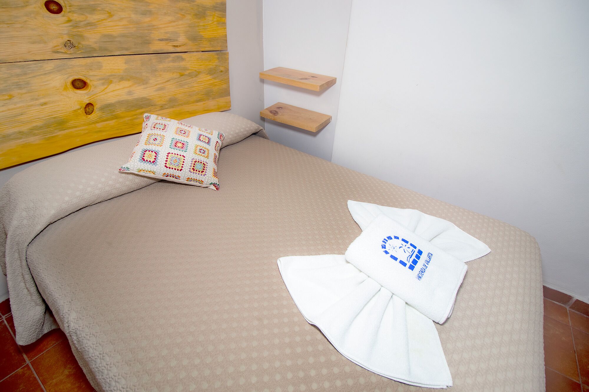 Cama bem arrumada com colcha bege, almofada colorida e toalha dobrada com um logotipo. Tem uma cabeceira de madeira e uma pequena prateleira acima da cama.