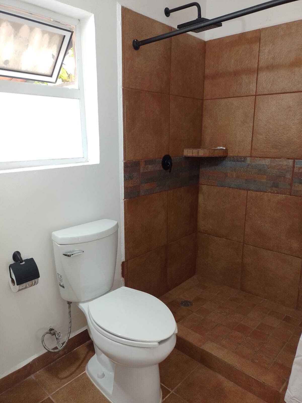 A bathroom with a white toilet, a shower area with brown tiles, and a window with blinds.