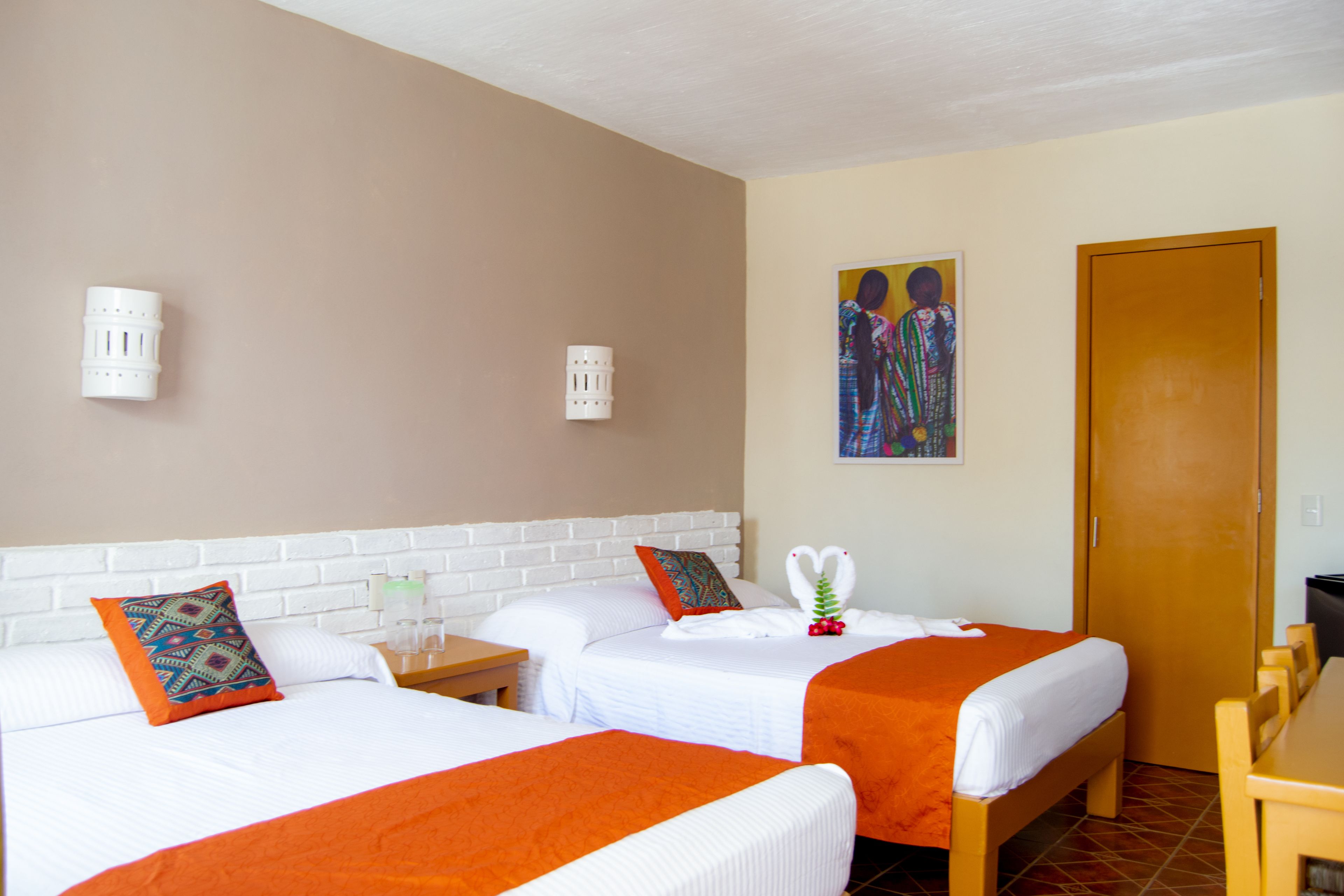 Habitación de hotel con dos camas, cada una con cubrecama naranja, ropa de cama blanca y almohadas decorativas. Hay un escritorio de madera con silla, una lámpara de pared y una puerta a la derecha.