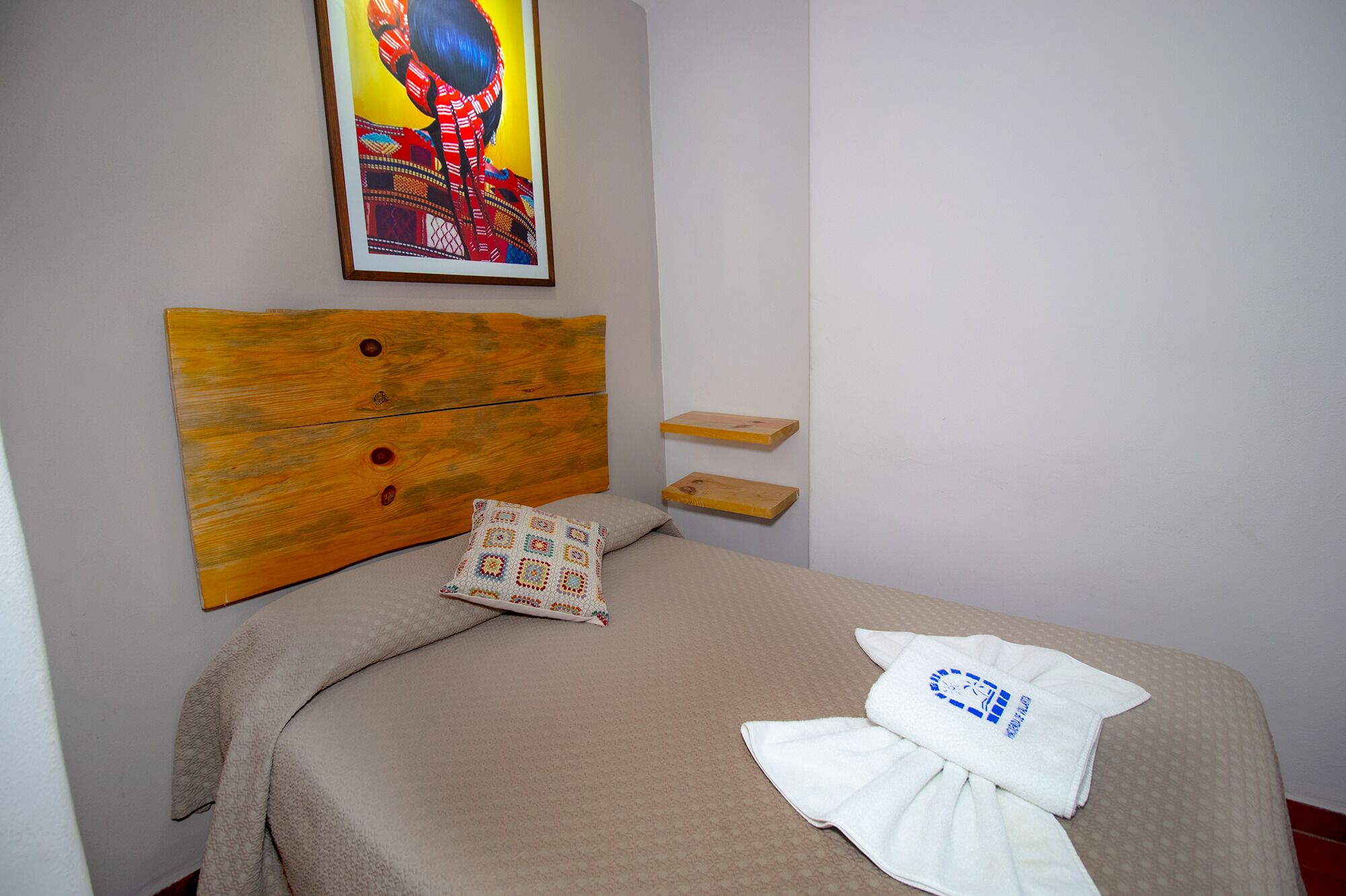Un lit bien fait, recouvert d’une couvre-lit beige, avec un oreiller blanc orné d’un logo bleu et une tête de lit en bois. Un tableau est accroché au mur, au-dessus du lit.