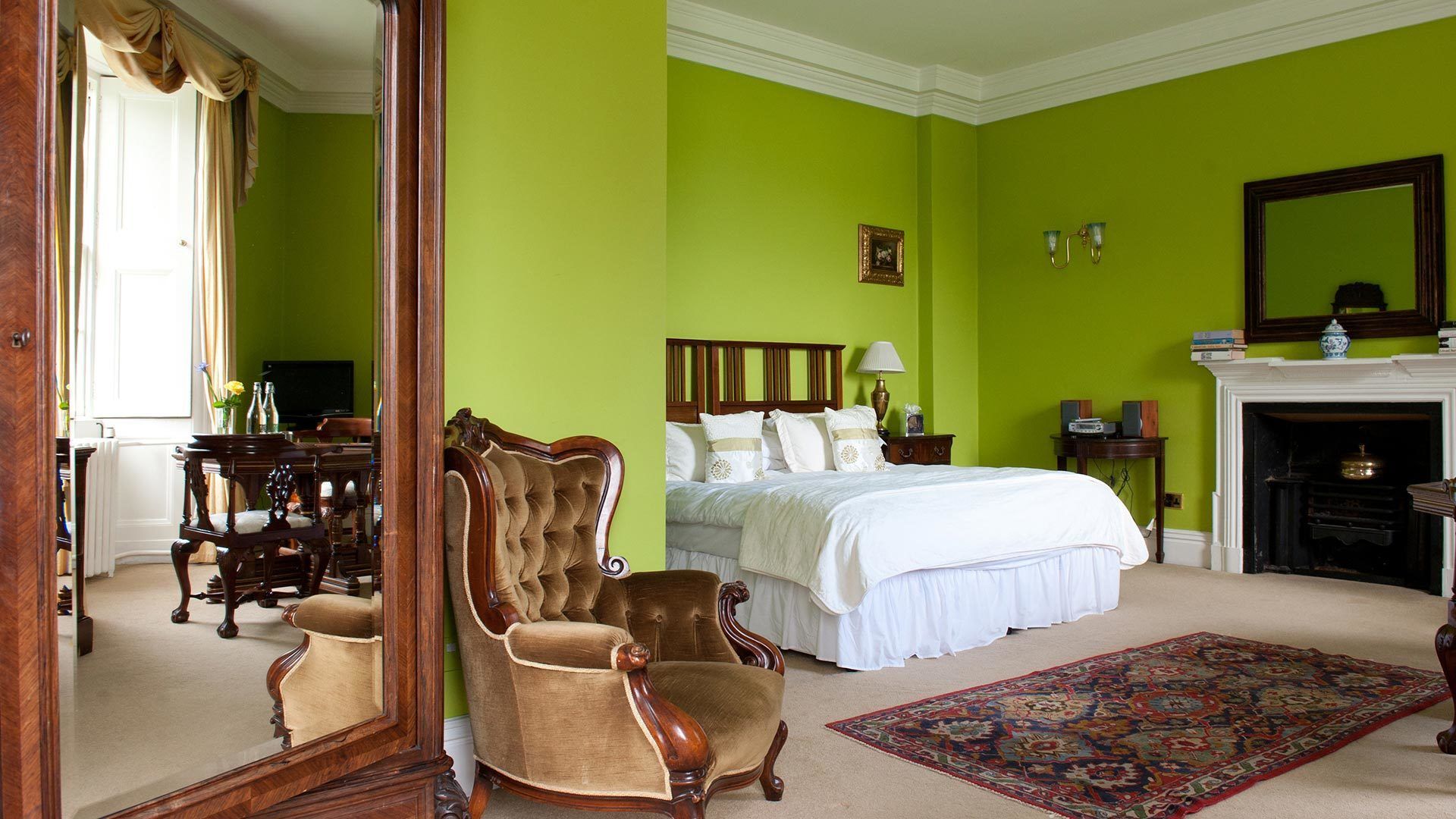 A bedroom with a large bed, a sitting chair, a fireplace, and a mirror.