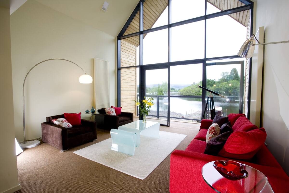 Una sala moderna con un sofá rojo, una mesa de centro de vidrio y vista a un lago a través de grandes ventanales.