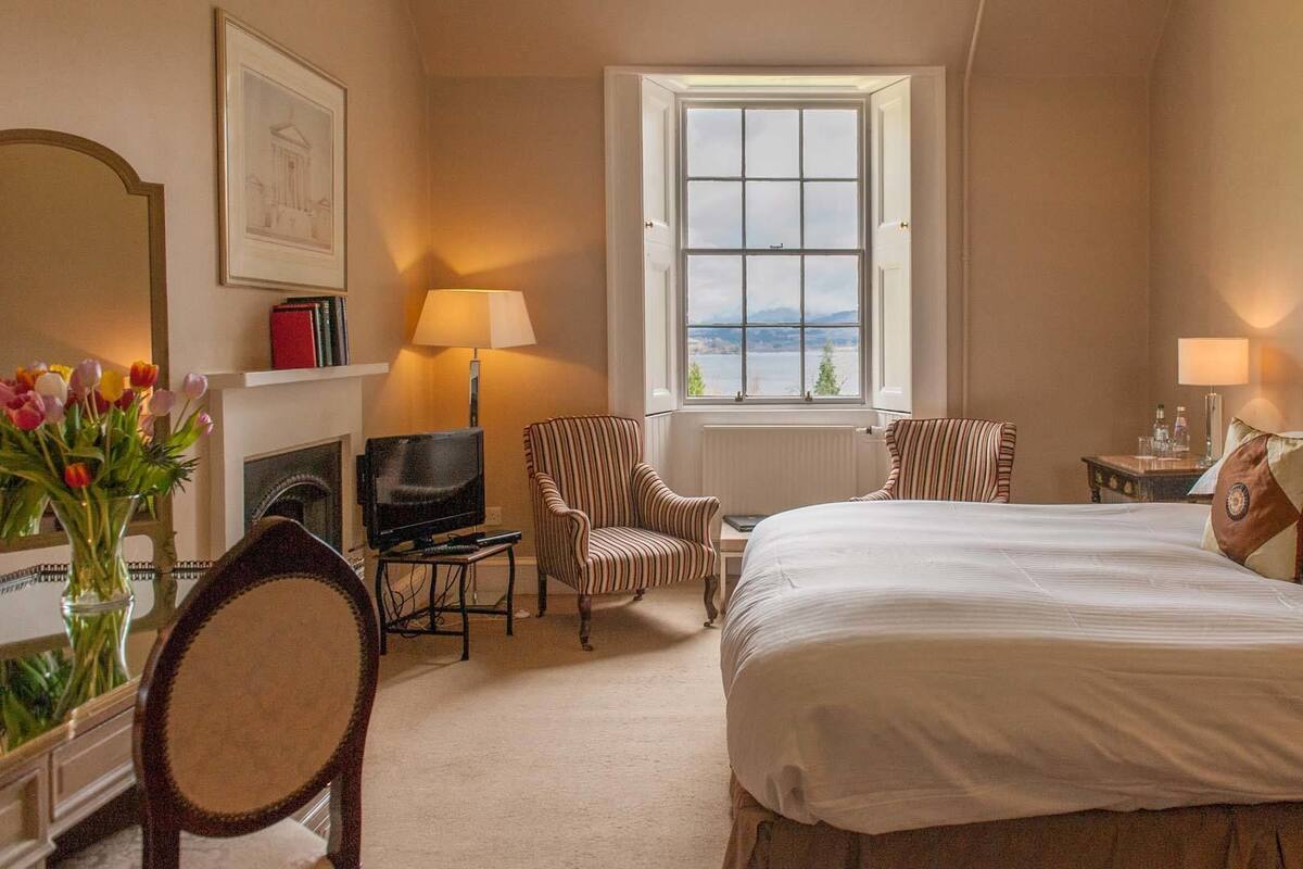 Habitación de hotel con una cama grande, dos sillones, un televisor, chimenea y ventana con vista al mar.