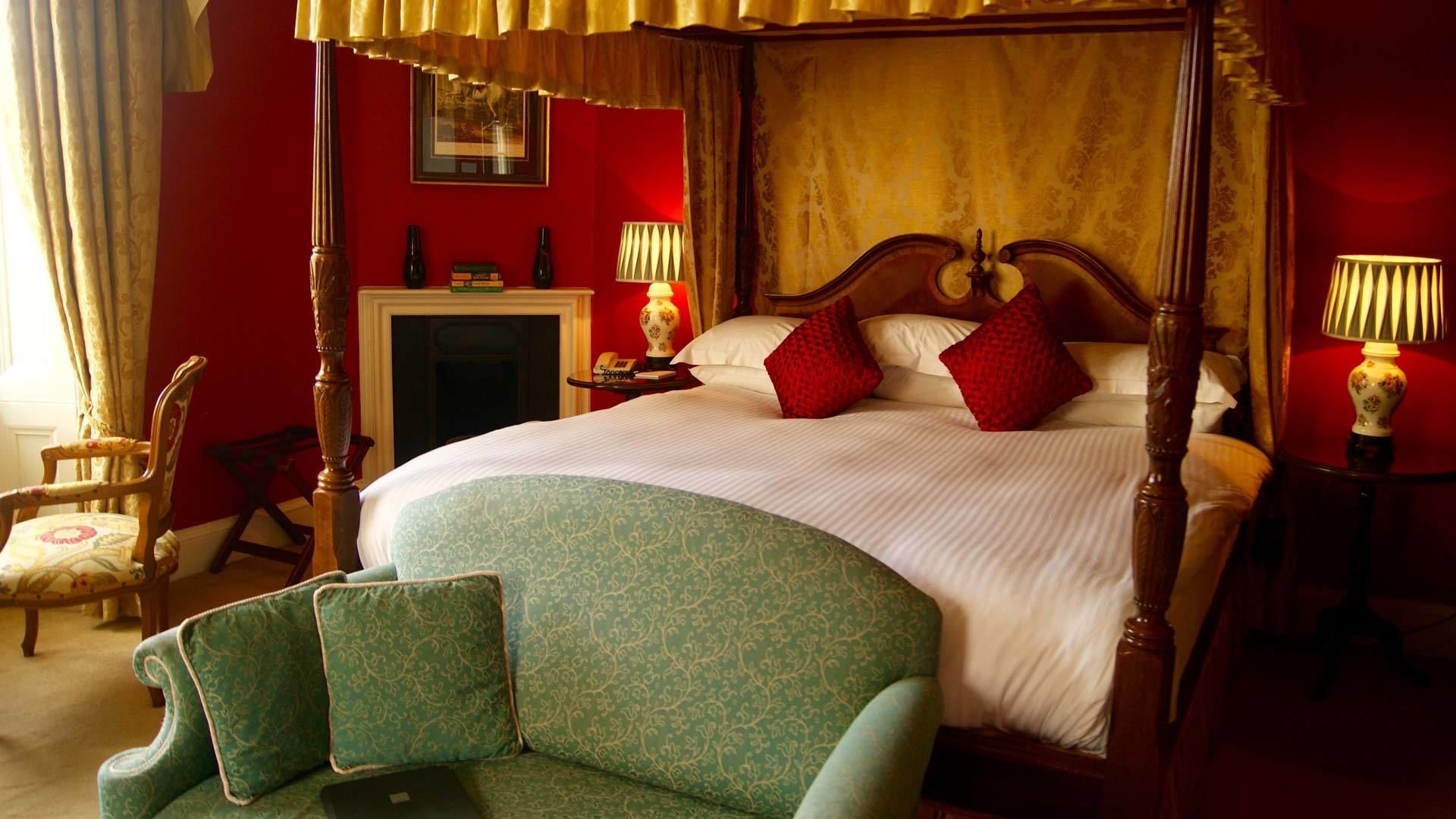 A four-poster bed with a red wall and a fireplace. There is a green armchair and a wooden chair.