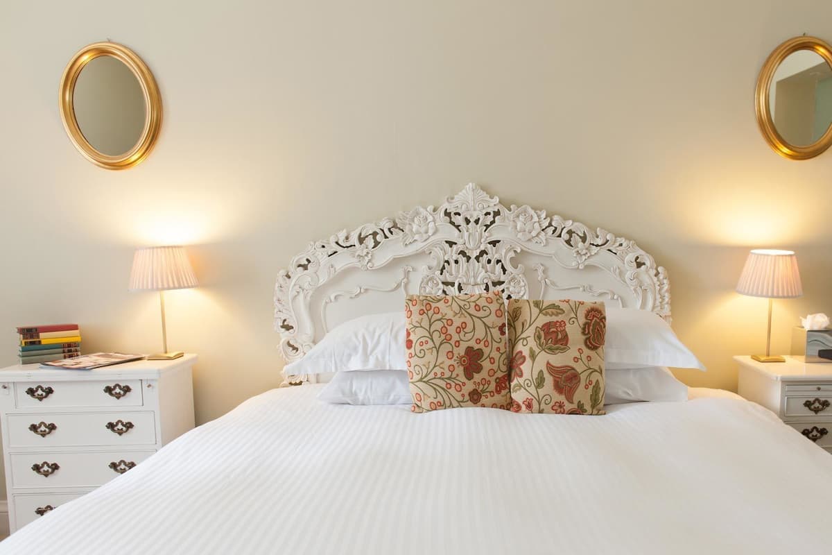 Una cama bien hecha con cabecera blanca y almohadas estampadas con motivos florales, flanqueada por mesitas de noche con lámparas y libros.