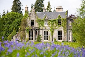 A historic stone building with ivy-covered walls and multiple windows, surrounded by lush greenery and purple flowers.