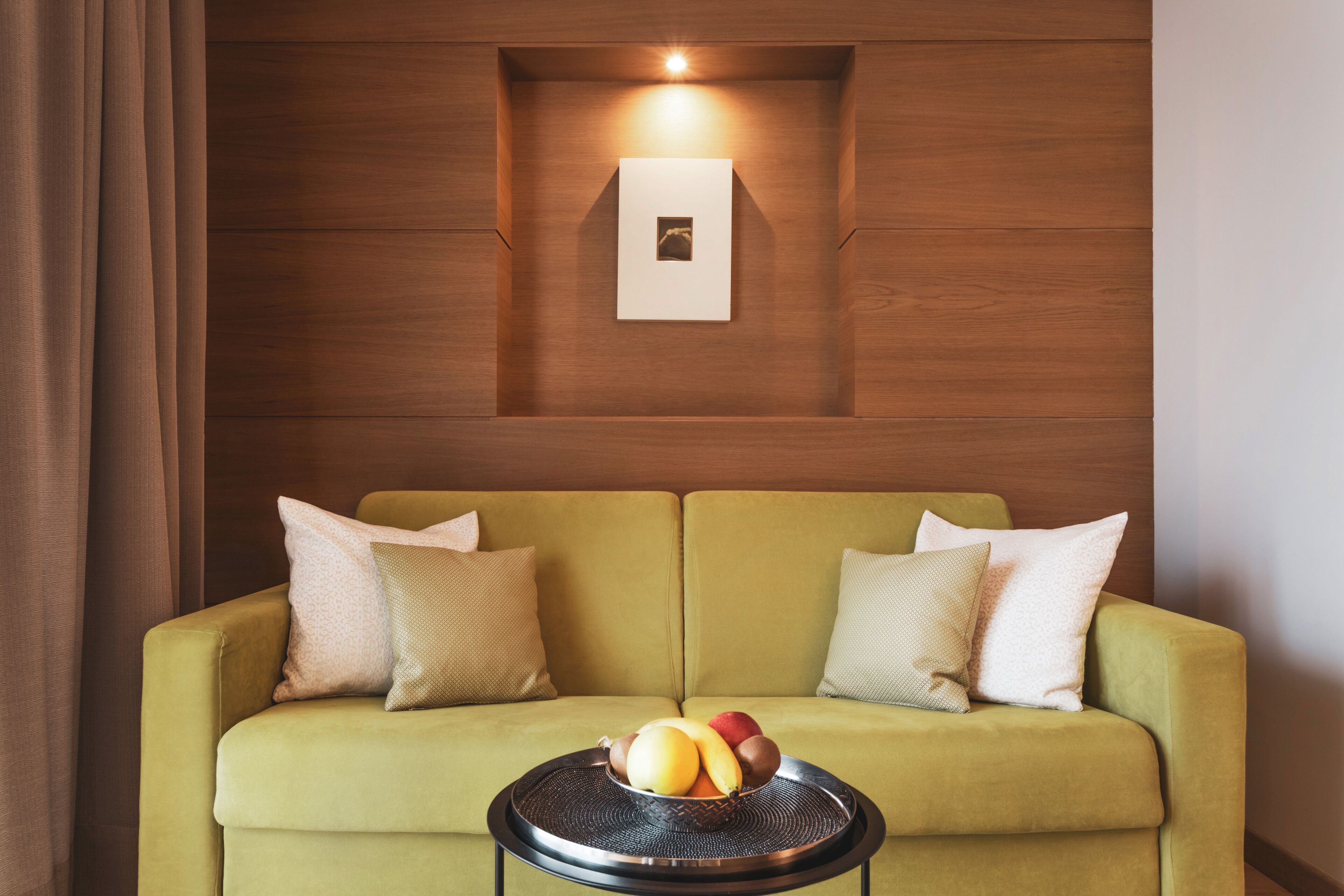 A green sofa with a fruit bowl on a round table, a wall-mounted picture, and a light fixture above the picture.