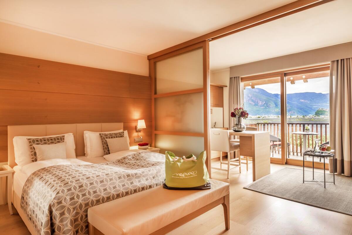 A hotel room with two beds, a balcony with a view of mountains, a desk, and a green bag on the bed.