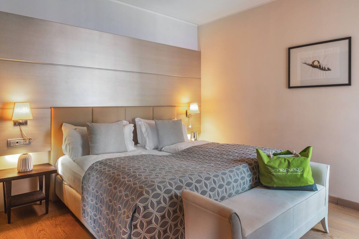 A hotel room with a large bed, bedside lamps, a green bag on a chair, and a framed picture on the wall.