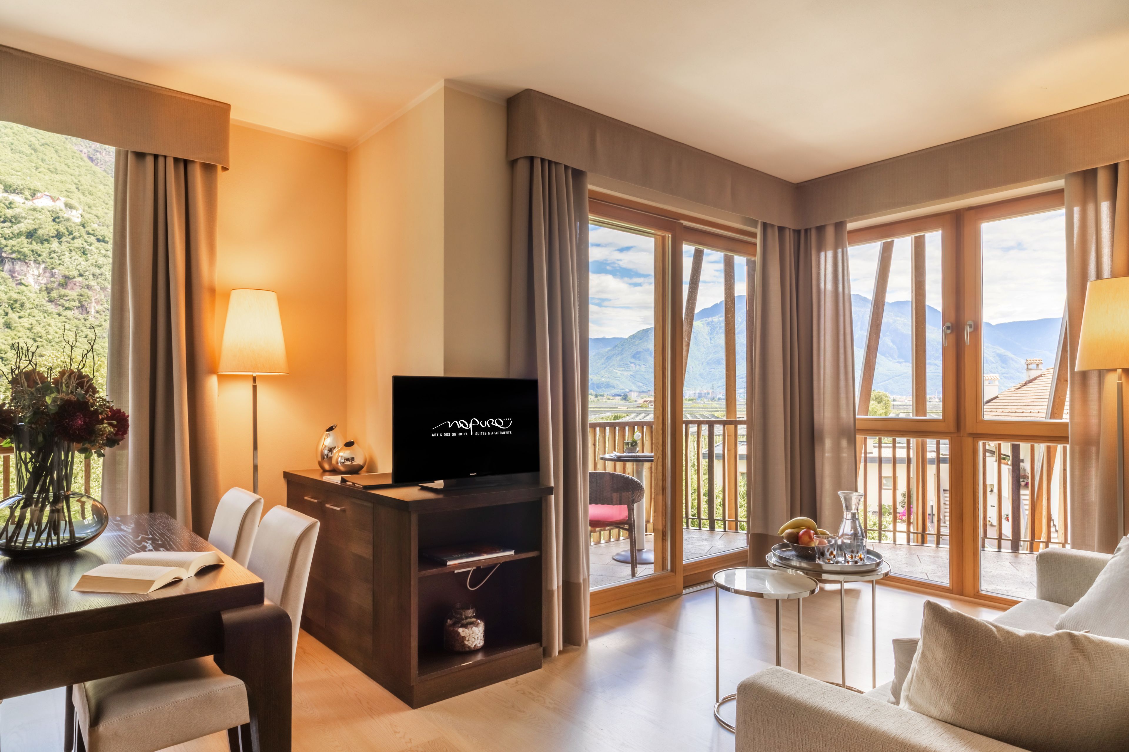 Habitación de hotel con zona de comedor, televisión y balcón con vistas a la montaña.