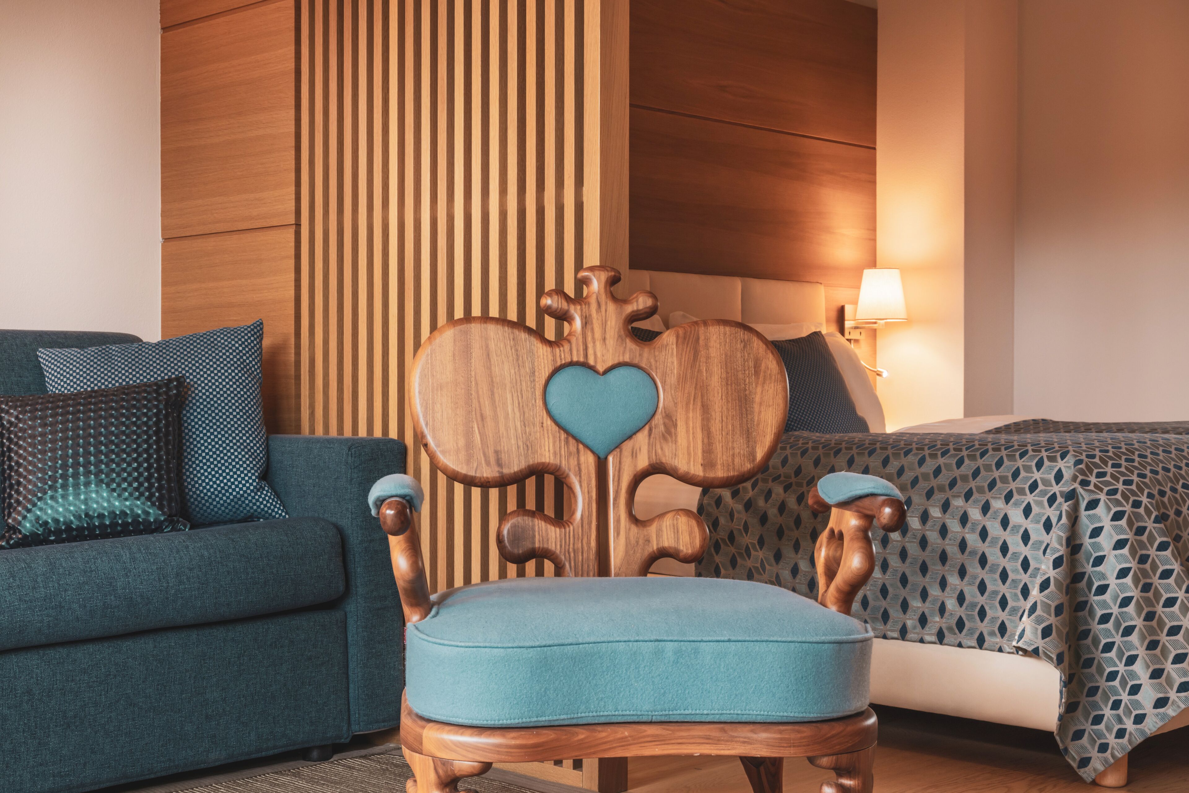 A room with a wooden headboard with a heart-shaped design, a blue upholstered chair, a blue sofa with patterned cushions, and a bed with a geometric patterned bedspread.