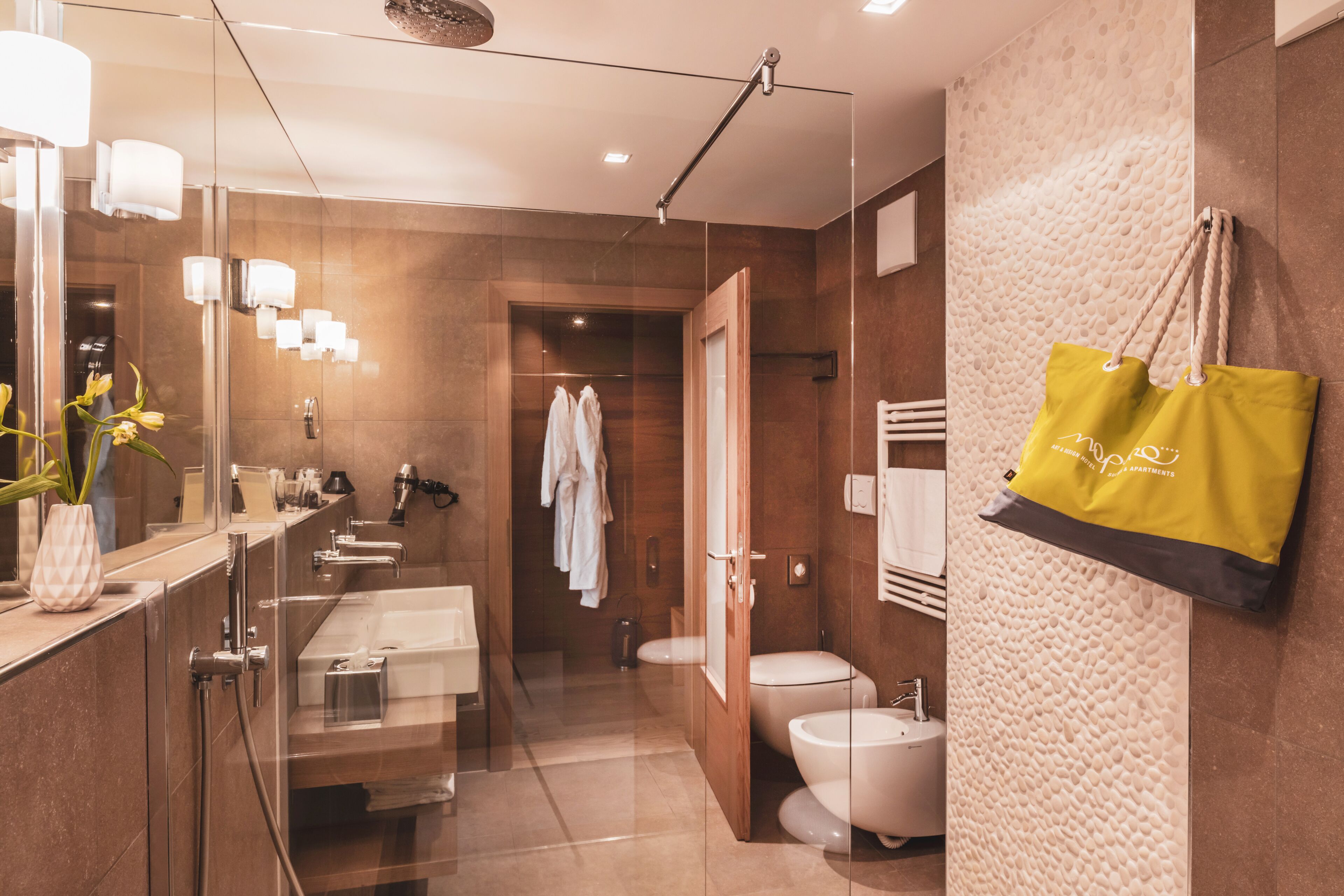 A modern bathroom with a glass shower enclosure, a large mirror, and a yellow tote bag hanging on the wall.