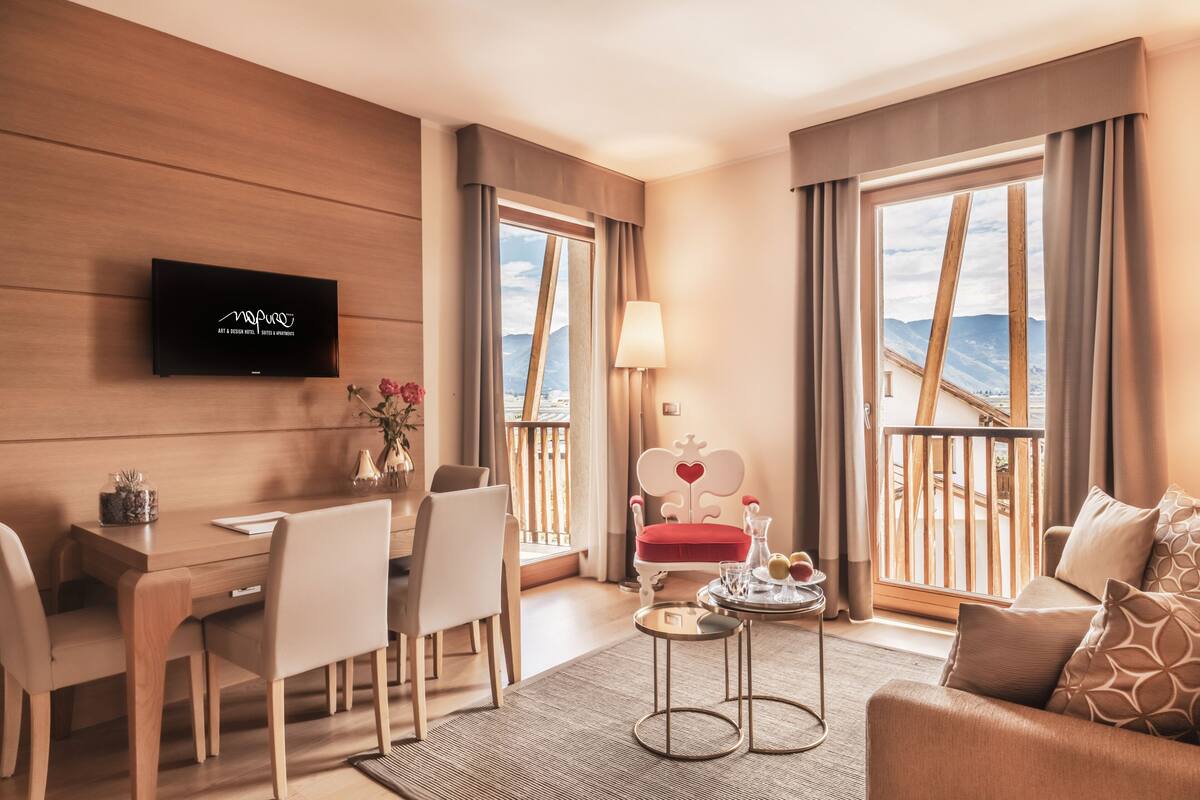 A modern hotel room with a wooden wall, a flat-screen TV, a dining area with a table and chairs, a red chair with a heart design, a sofa, and large windows with a view of mountains.