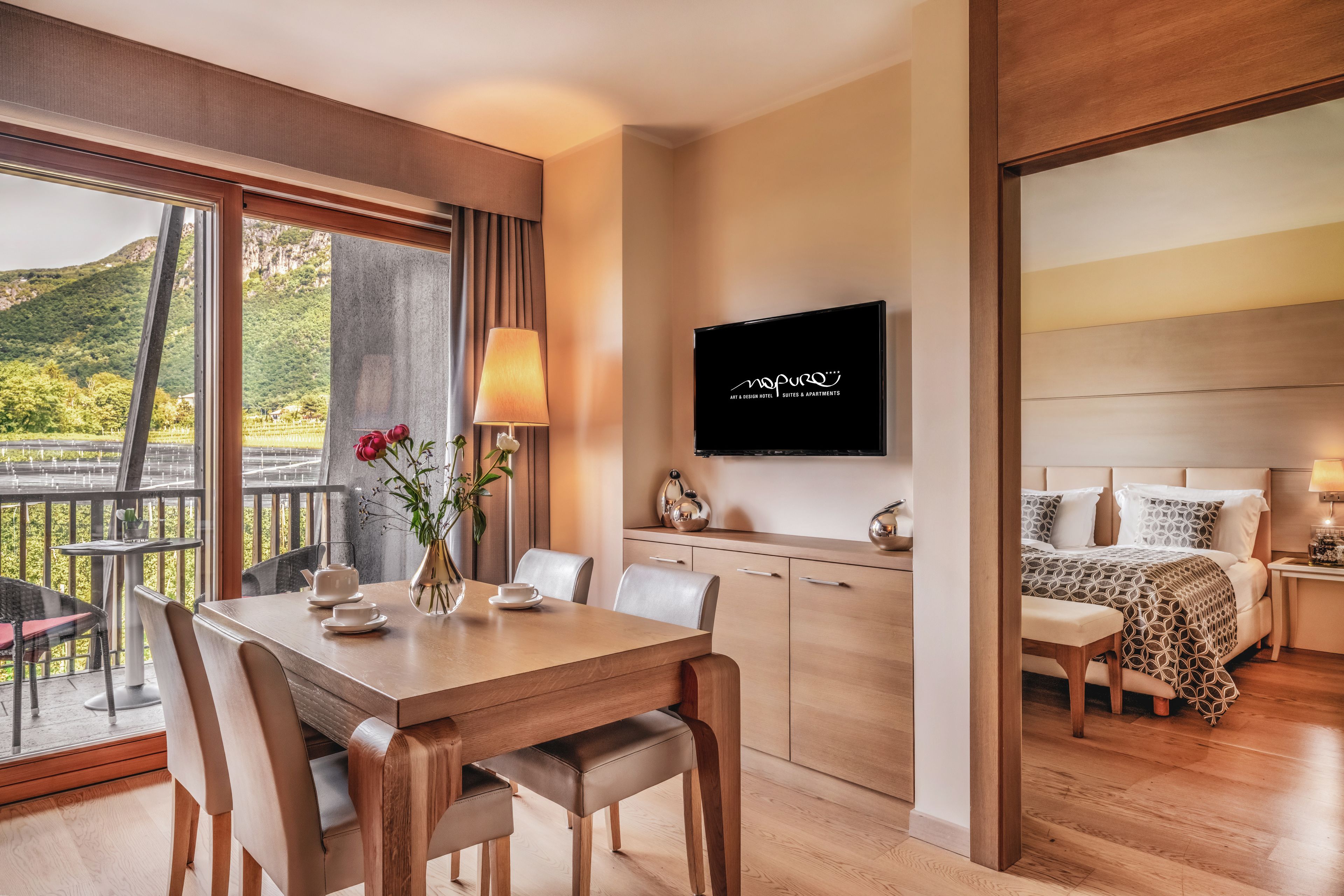 A modern hotel room with a dining area, a television, and a balcony with a view of greenery.