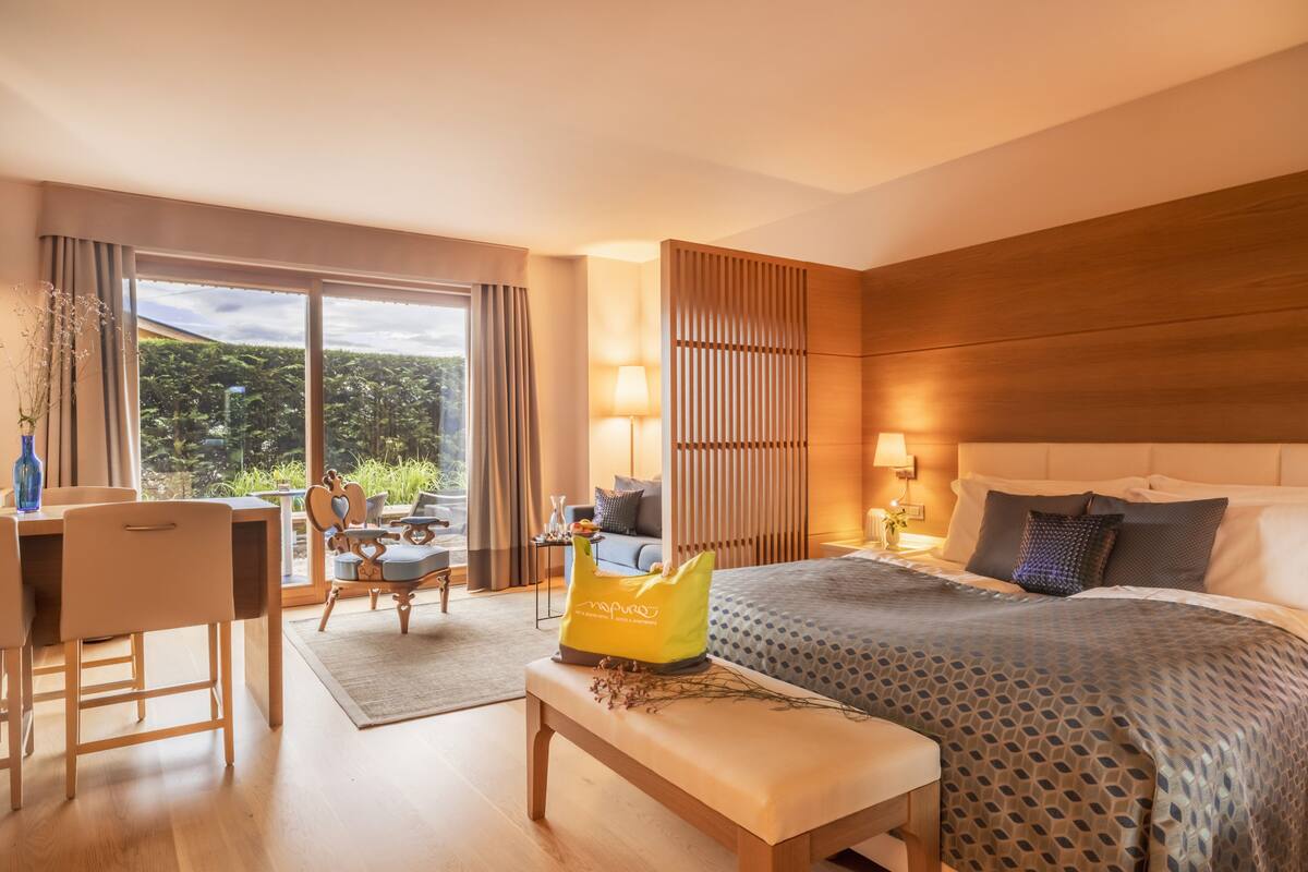 A hotel room with a bed, a desk, a chair, a small table with a lamp, and a view of greenery outside.