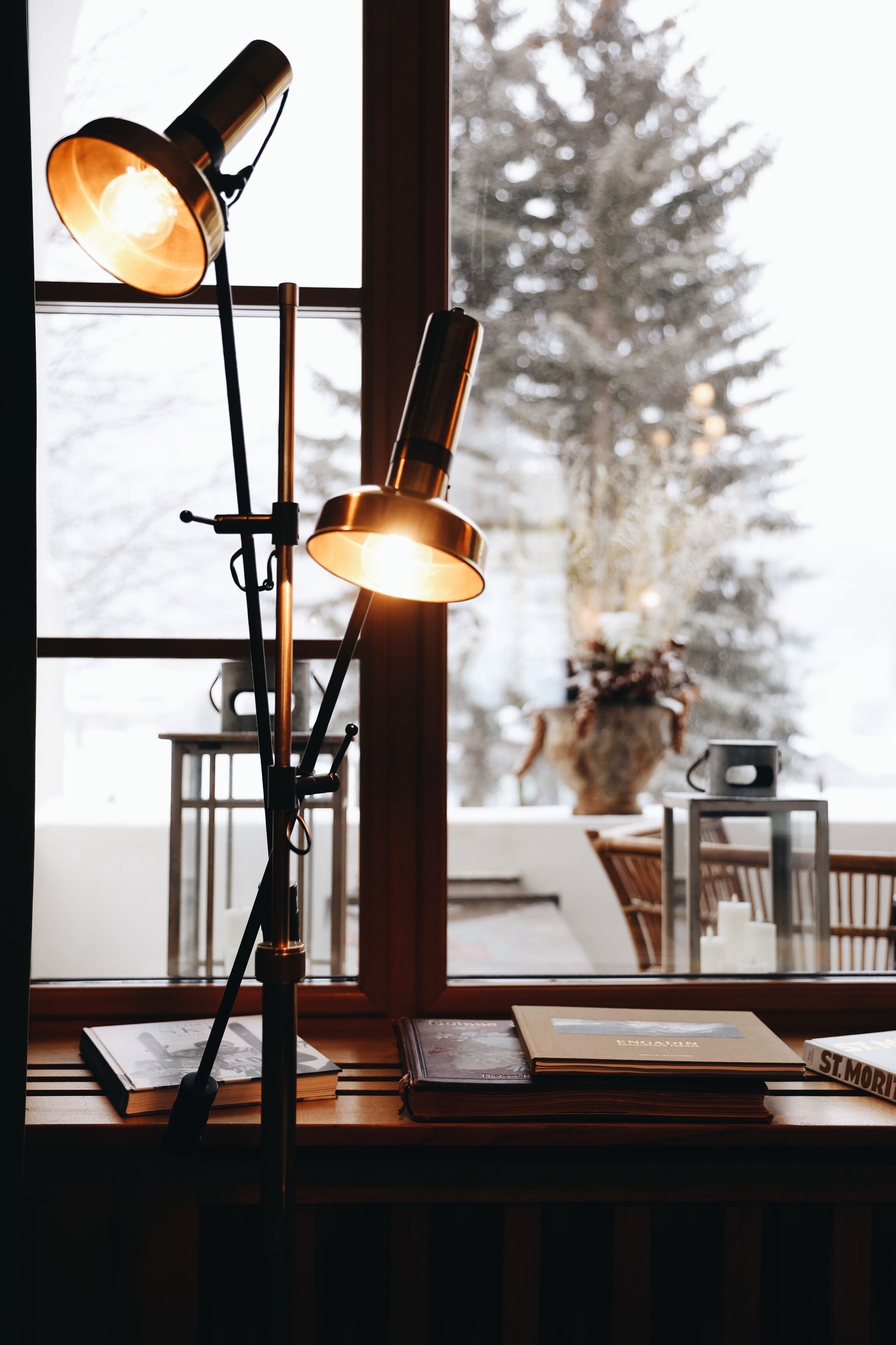 A desk with books and a lamp near a window with a view of a snow-covered tree.
