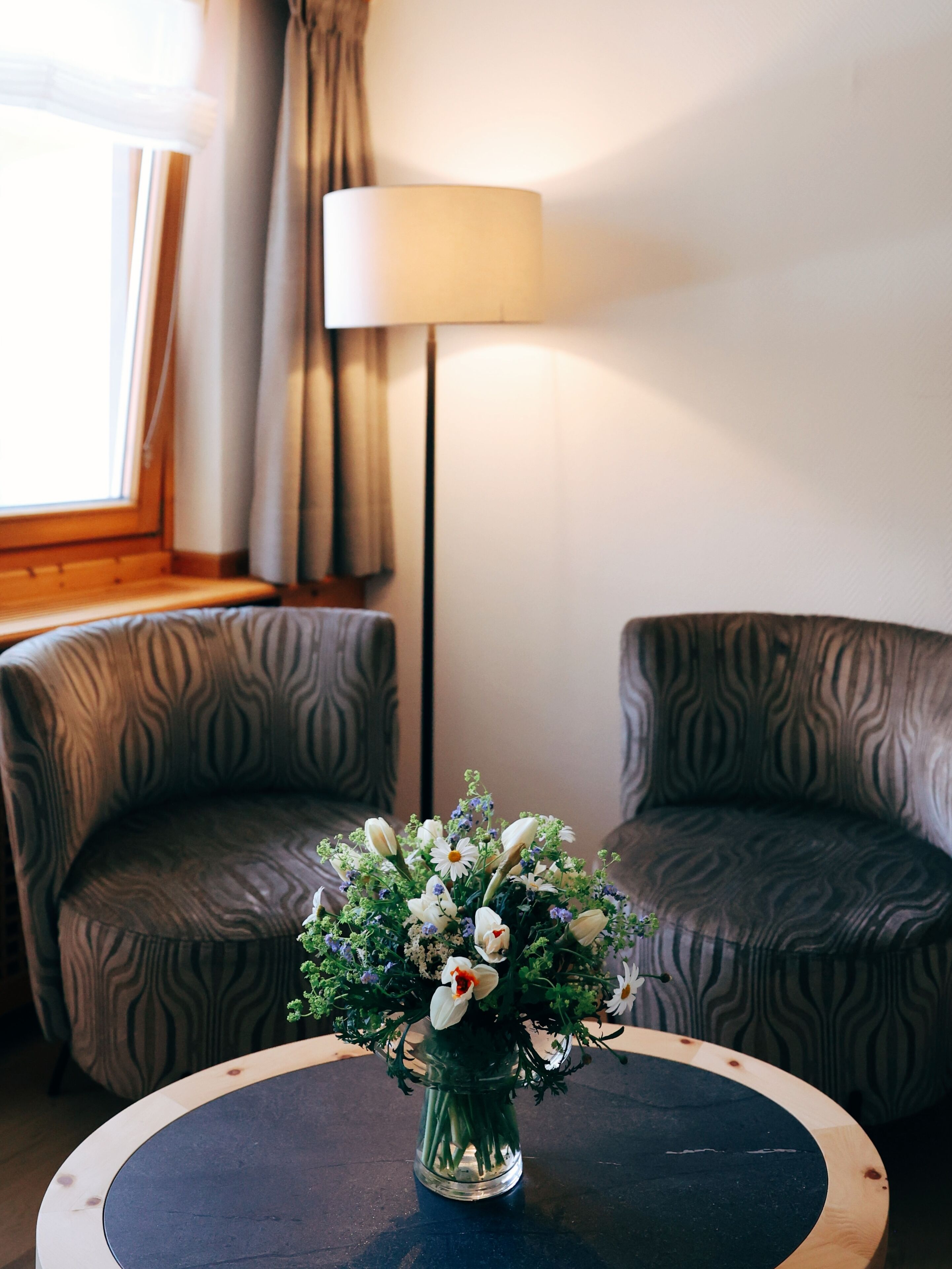 A room with two patterned armchairs, a round table with a vase of flowers, a lamp, and a window with curtains.