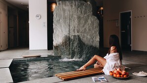 Una persona relajándose junto a una fuente de piedra en el vestíbulo de un hotel, con un tablón de madera, una cesta de fruta y una revista en el suelo.