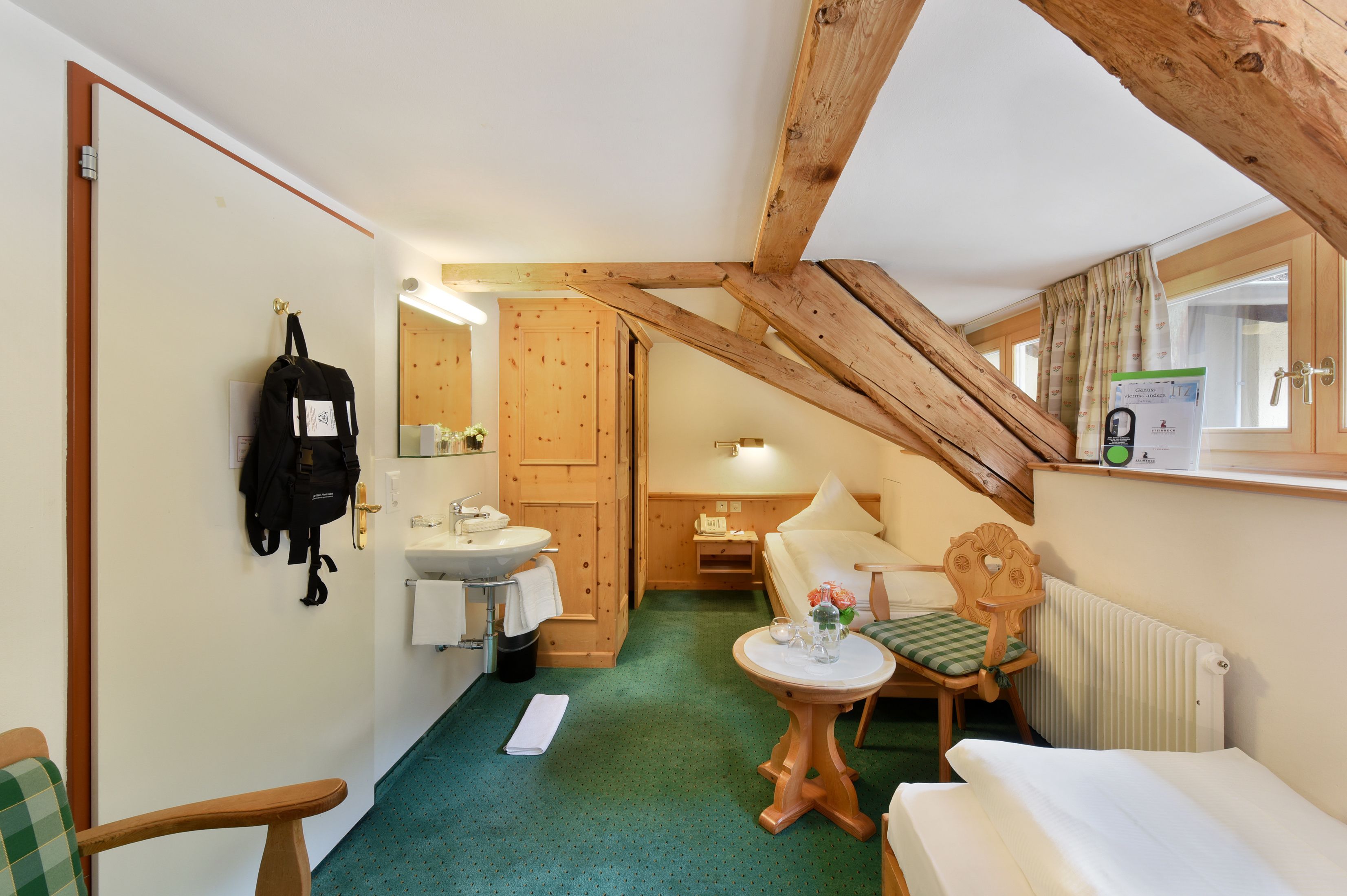 A compact hotel room with a wooden ceiling, a small bathroom with a sink and mirror, a single bed, a chair with a table, and a window with curtains.