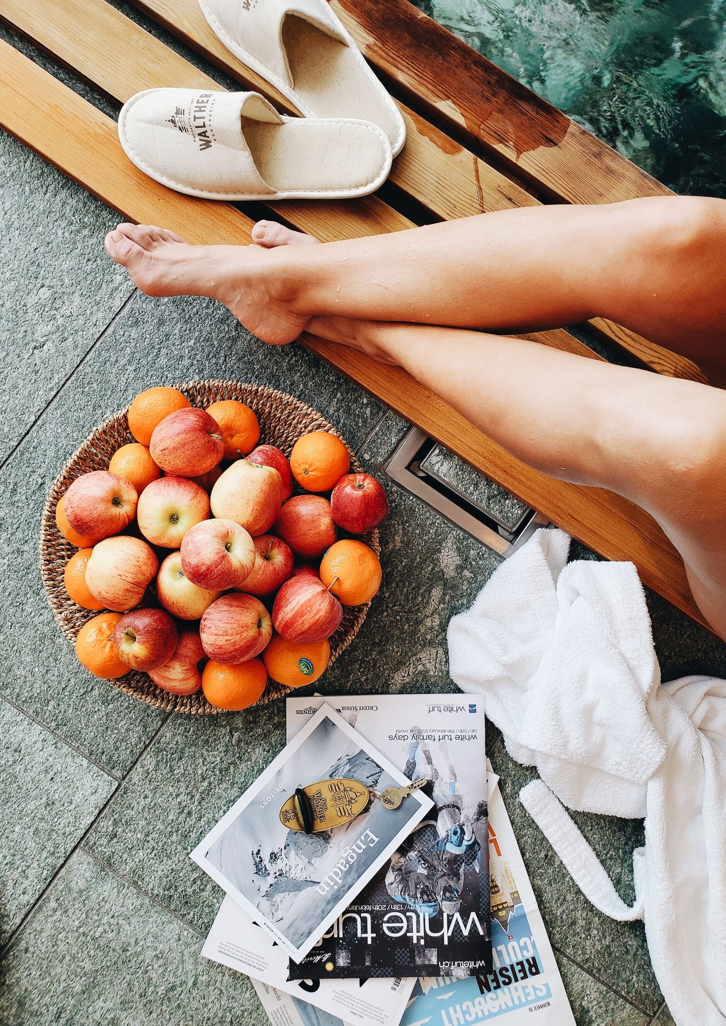 Pies descansando sobre un banco de madera, una cesta de manzanas y naranjas, y algunas revistas en el suelo.