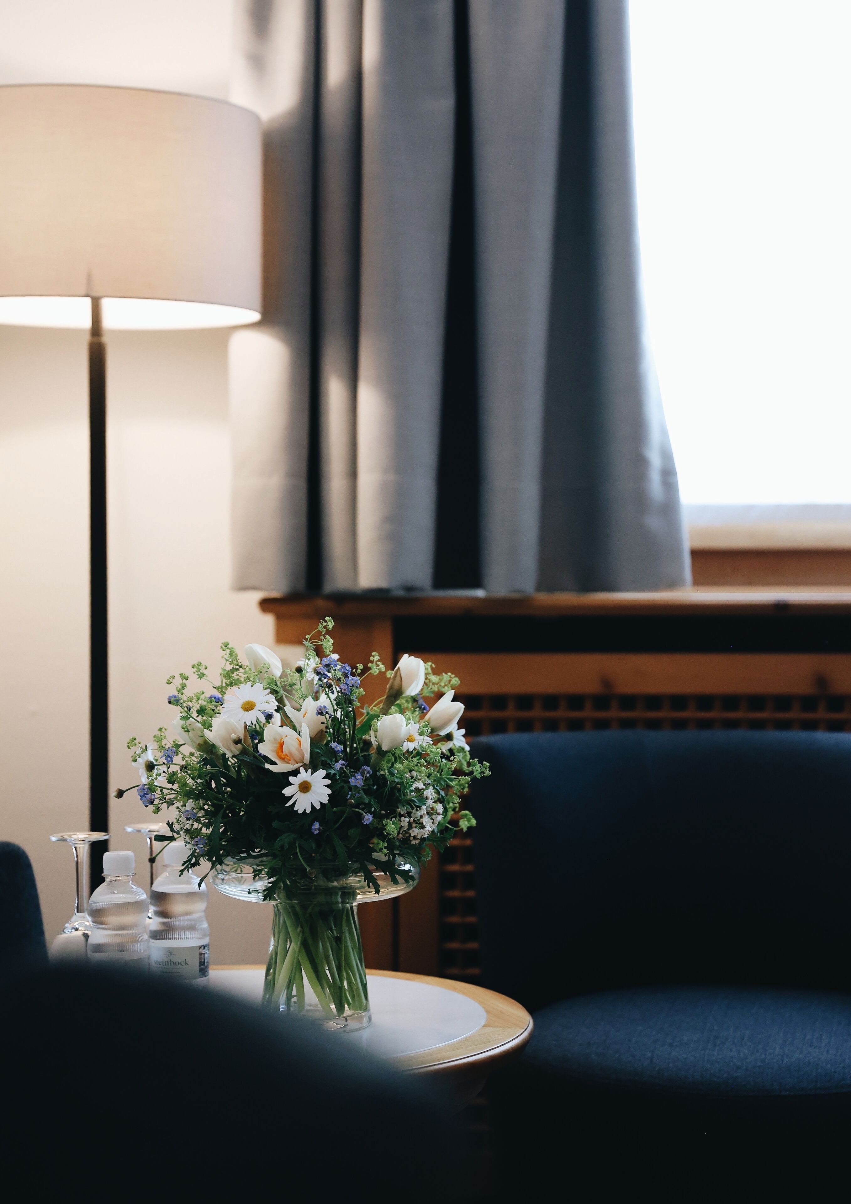 A hotel room with a dark blue sofa, a round table with a vase of flowers, a lamp, and a window with curtains.