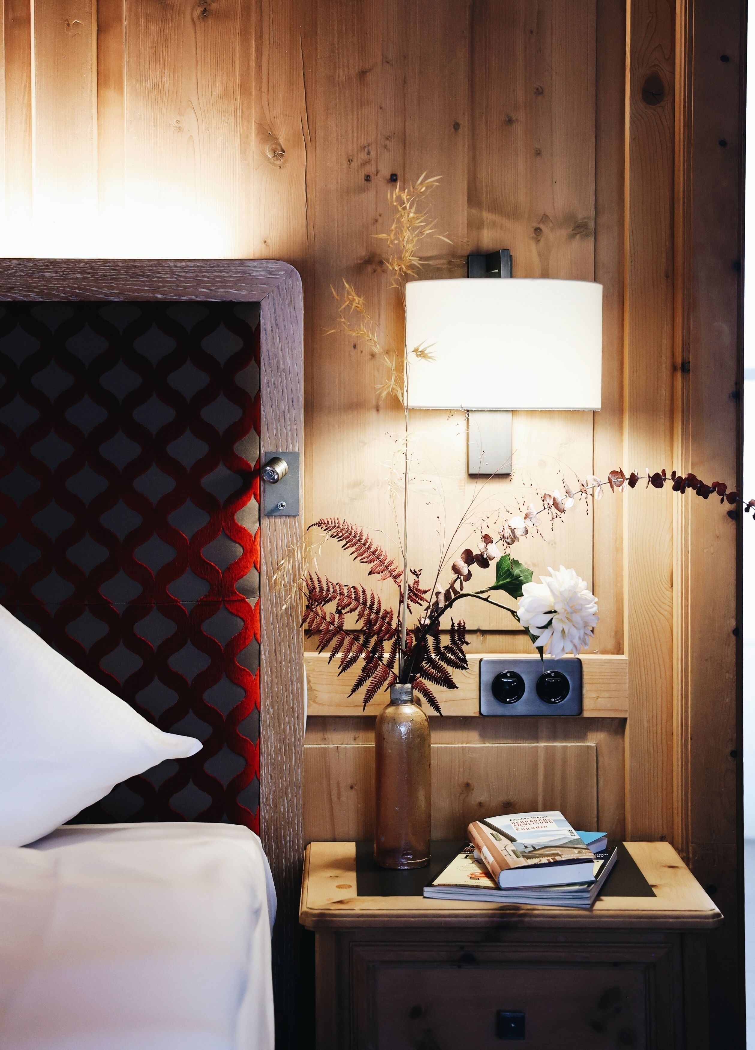 Una habitación con paneles de madera, una cama, una mesita de noche con lámpara, un jarrón con flores y una radio.