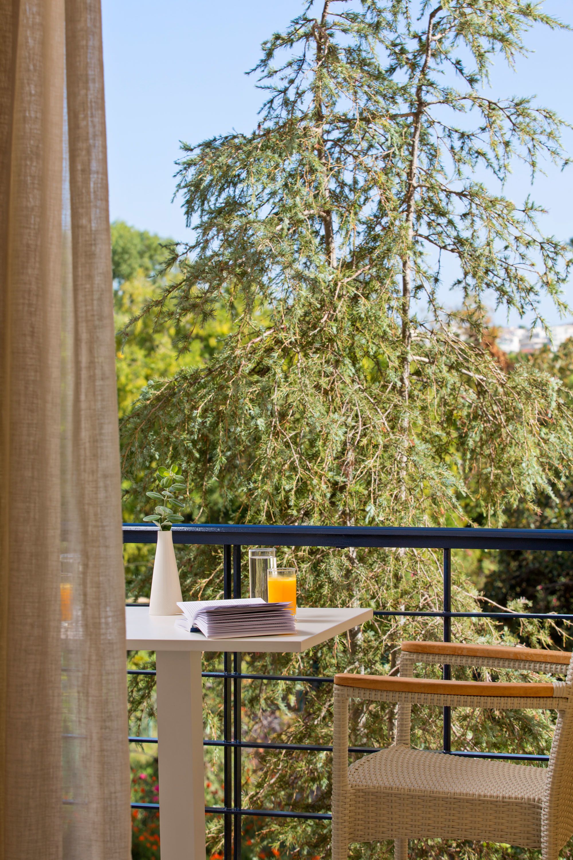 A balcony with a wicker chair, a small table with a glass of orange juice, and a view of a tree.
