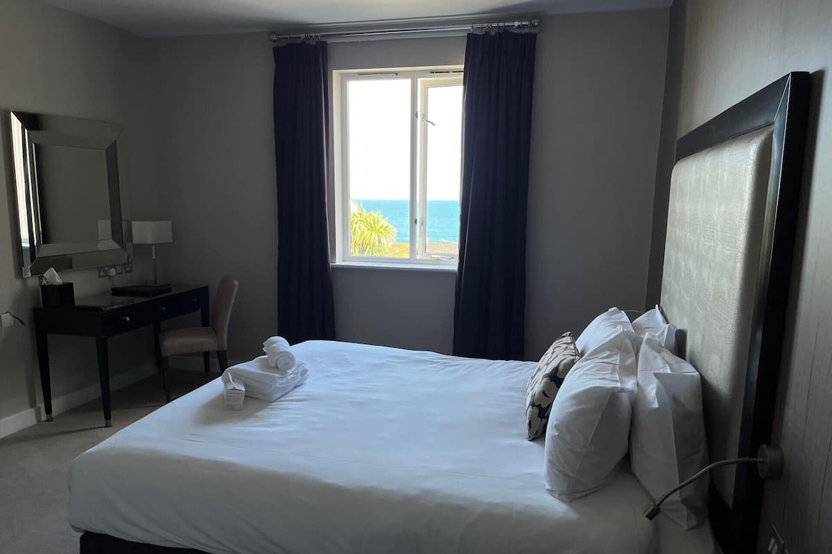 A hotel room with a bed, a desk, a chair, a window with curtains, and a view of the sea.