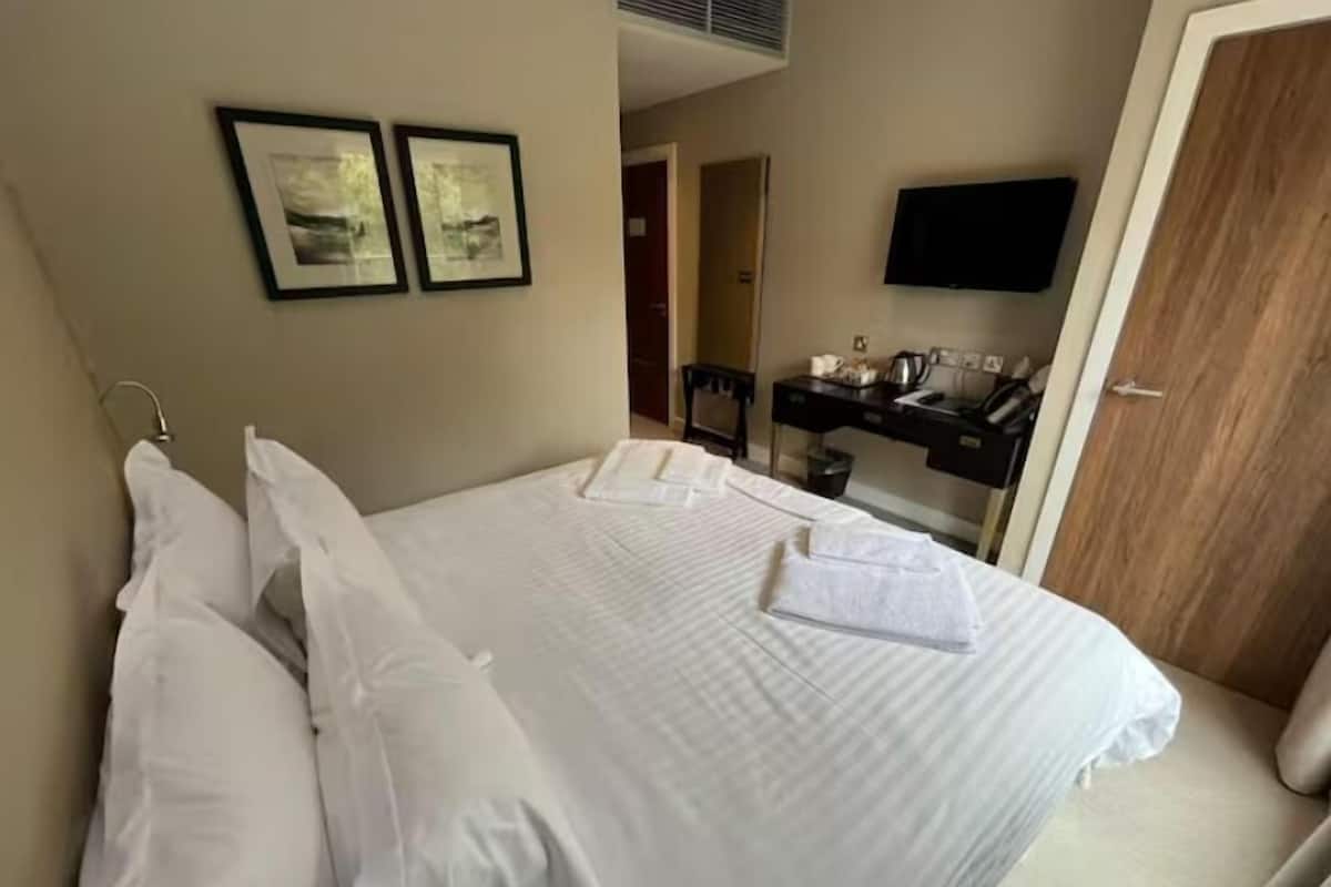 A hotel room with a bed, white linens, a desk with a flat-screen TV, and two framed pictures on the wall.