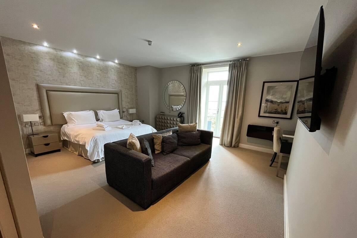 A modern hotel room with a large bed, a sofa, a TV, and a bathroom visible through the door.