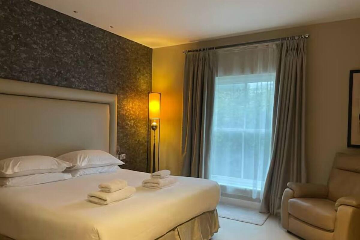 A hotel room with a large bed, a headboard, a bedside table, a lamp, a window with curtains, and a chair.