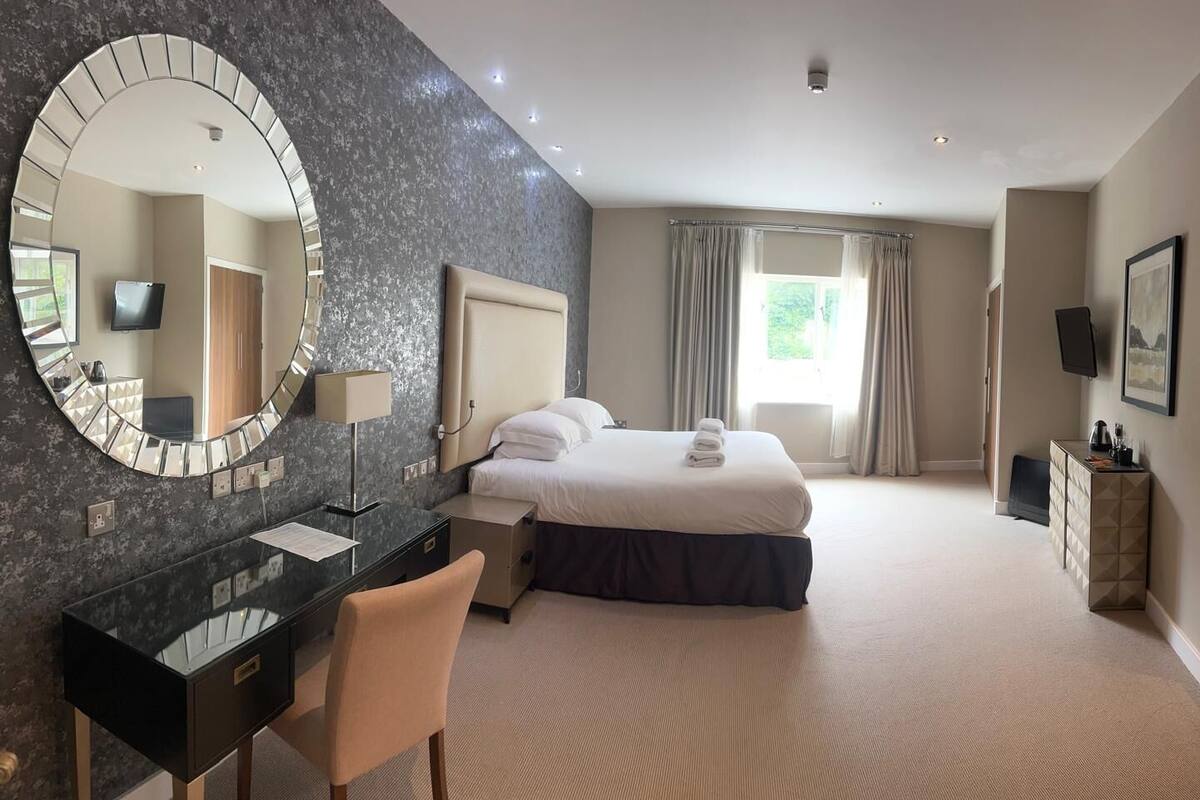 A hotel room with a bed, a desk with a chair, a large mirror, and a television.