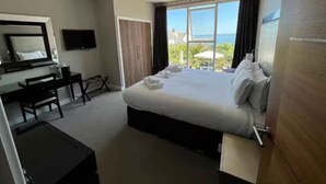A hotel room with a bed, a desk, a chair, a TV, and a large window with a view of the sea. - The Llawnroc  (St Austell)