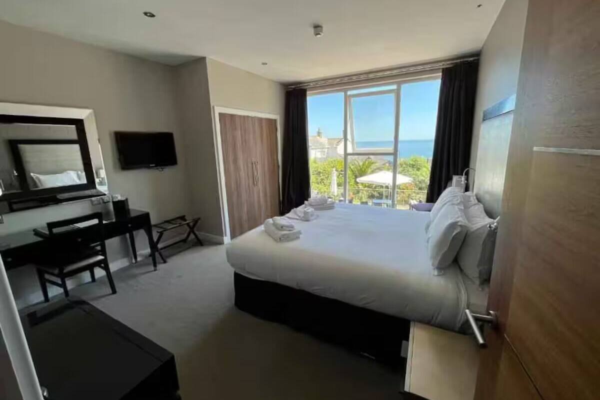 A hotel room with a bed, a desk, a chair, a TV, and a large window with a view of the sea.