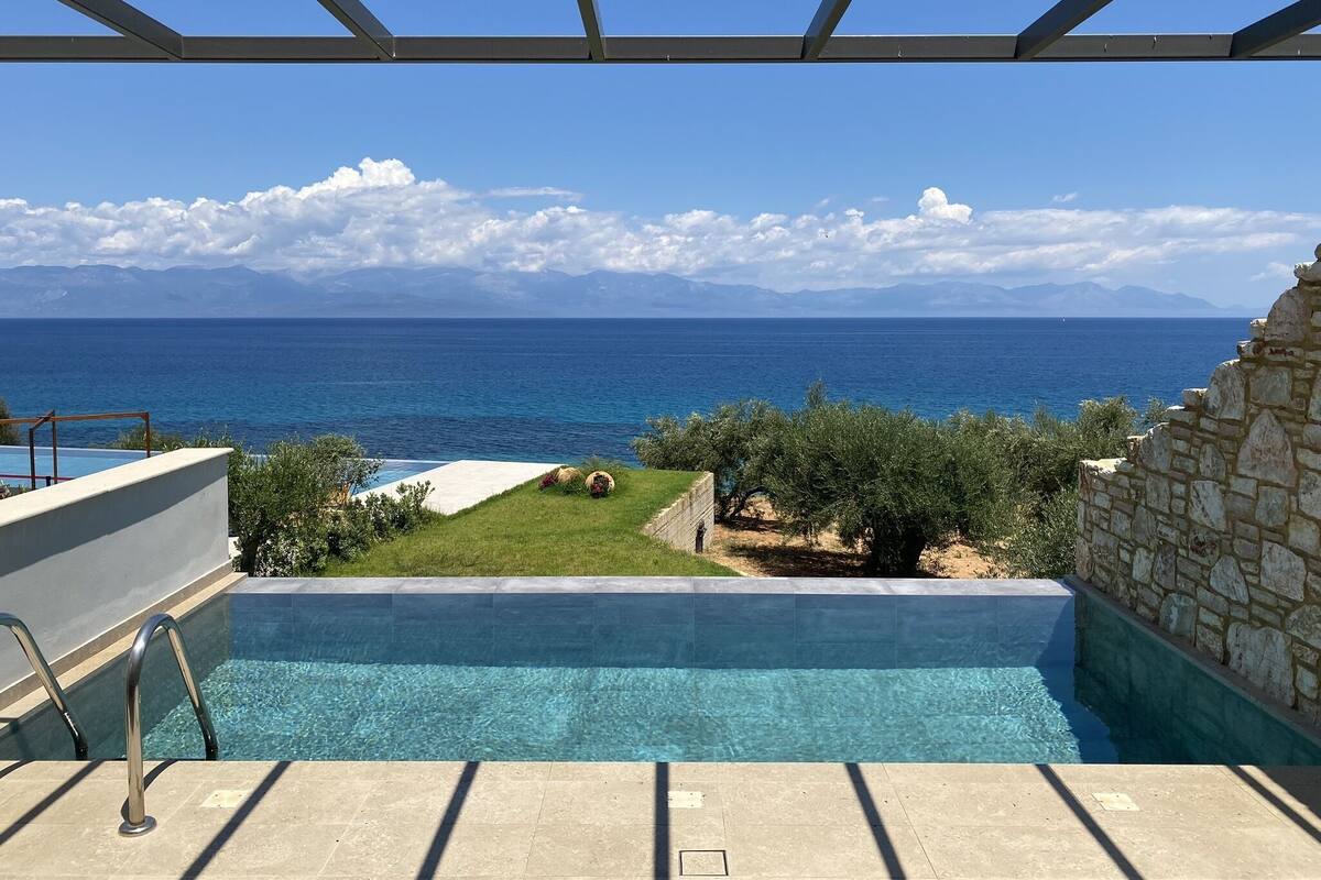 A pool with clear blue water, a stone wall, and a view of the sea and mountains in the background.