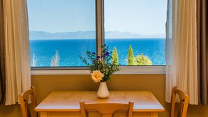 A room with a wooden table and chairs, a vase with flowers, and a view of the sea and mountains through a window with curtains. - Colonides Beach Hotel (Pylos-Nestoras)