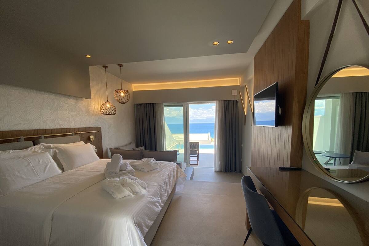 A hotel room with a large bed, a desk, a chair, a TV, and a balcony with a view of the ocean.
