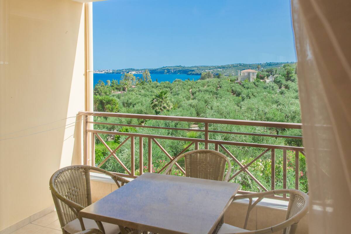 A balcony with wicker furniture and a table, overlooking a coastal landscape with greenery and a clear blue sky.