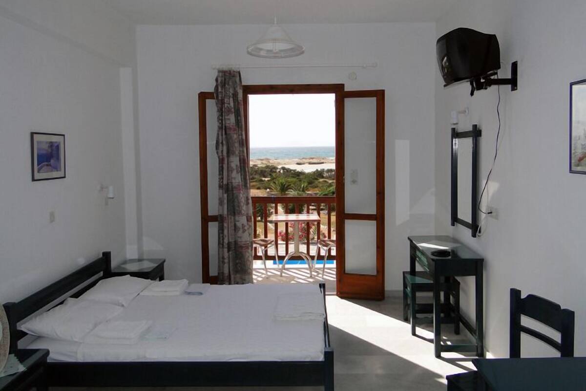 A bedroom with a bed, a desk, a chair, a TV, and a balcony with a view of the beach.