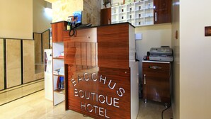 Un mostrador de recepción con detalles de madera y el nombre "BACHUS BOUTIQUE HOTEL". Se observa un pilar de piedra, una unidad de aire acondicionado y un pequeño escritorio con una impresora.
